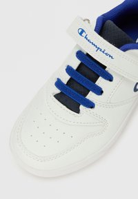 Baskets Champion blanches avec lacets bleus, langue marine et sangle Velcro arborant le logo Champion.