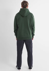 Sweat à capuche vert foncé avec une capuche, fabriqué en tissu doux. Présente des poignets et un ourlet côtelés, ainsi qu'un logo discret sur l'épaule. Assorti à un pantalon foncé.