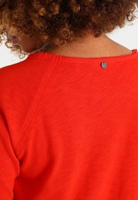 Rode T-shirt met korte mouwen en een ronde halslijn. Voorzien van een subtiele onafgewerkte rand en een kleine metalen accent nabij de achterkant van de halslijn. Zacht, gestructureerd materiaal.