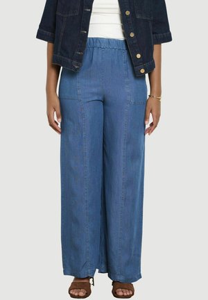 Kobieta ubrana w wysokie, szerokie, niebieskie dżinsy, biały top, krótką ciemną kurtkę dżinsową i brązowe sandały z odkrytymi palcami.