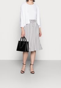 Donna che indossa un top bianco con cardigan abbinato, una gonna midi grigio e bianca a righe, sandali con tacco neri e tiene una borsa nera.