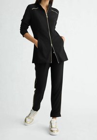 Czarny strój sportowy składający się z kurtki na zamek błyskawiczny ze złotymi akcentami oraz obcisłych spodni z bocznymi suwakami, zestawiony ze złotymi, akcentowanymi sneakersami.