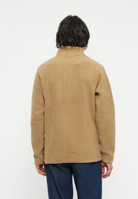 Fleece pullover in beige, met een hoge kraag, gestructureerd oppervlak en lange mouwen. De achterkant heeft een ontspannen pasvorm zonder zichtbare hardware.