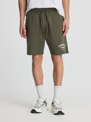 Uomo che indossa pantaloncini verdi oliva con coulisse e stampa bianca "Authentic Co", calzini bianchi alla caviglia, scarpe da corsa bianche e grigie, in piedi davanti a uno sfondo neutro.