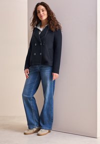 Navy peacoat-tyylinen takki, jossa on kaksiriviset napit ja huppu, yhdistettynä sinisiin leveälahkeisiin farkkuihin ja vaaleisiin lenkkareihin.