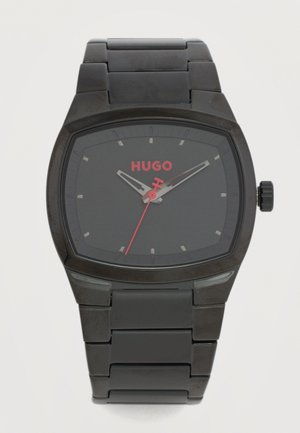 Fekete fém karóra téglalap alakú számlappal, ezüst óraszámozással, piros másodpercmutatóval és piros "HUGO" logóval a számlapon.