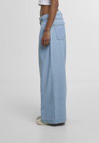 Light blue wide-leg jeans made of denim. Features a high waist, visible stitching, and two back pockets. Worn with light-colored shoes.