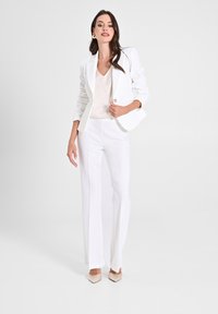 Blazer bianco con un solo bottone, abbinato a un top beige chiaro e pantaloni bianchi a gamba larga. Tacchi beige a punta completano l'outfit.