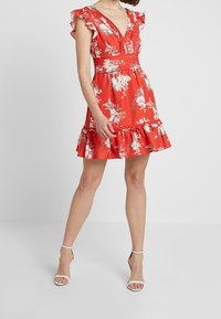 Červené květinové šaty s volánkovými rukávy, hlubokým výstřihem do V, přiléhavým pasem a rozšířeným lemem. Vyrobené z lehké látky se vzorem bílých květin.