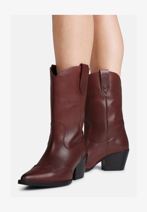 Pernas a usar botas de couro castanhas estilo ocidental até à altura da panturrilha, com saltos bloco e abas de puxar, sobre um fundo branco.