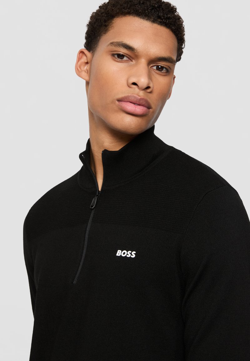 Schwarzer Zip-Kragen-Pullover aus gestricktem Stoff, mit einem strukturierten oberen Einsatz und einem weißen "BOSS"-Logo auf der linken Brust.