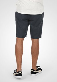 Shorts bleu foncé avec une texture lisse, coupe droite et deux poches latérales. Portés avec des baskets noires et blanches avec un accent doré.