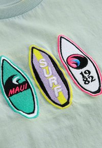 Šviesiai žalia medžiaga, puošta trimis siuvinėtą banglentes: turkio spalvos su užrašu "MAUI", violetinės su užrašu "SURF" ir baltos su užrašu "1982". Ryškūs akcentai.