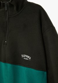 Pulóver de forro polar en verde oscuro y negro, con un alto cuello, cremallera de un cuarto y un logotipo bordado en blanco que dice "California". Textura suave.