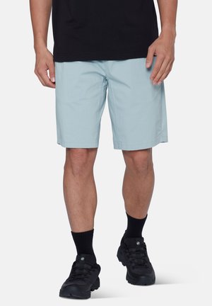 Homme portant un short bleu clair arrivant aux genoux, des chaussettes noires, des chaussures de randonnée noires et une chemise noire, debout sur un fond blanc uni.