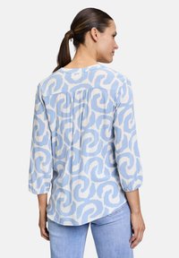 Blusa azzurro chiaro con motivo a spirale crema, maniche a tre quarti, dettaglio a pieghe sul retro e orlo arrotondato. Sotto sono indossati pantaloni in denim.