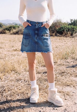 Una persona está de pie en un campo seco, con una prenda superior blanca, una falda vaquera deshilachada, calcetines blancos y botas de invierno.