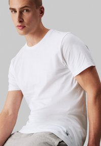 T-shirt en coton blanc avec un col rond, des manches courtes et un logo discret en bas, associé à un bas gris. Texture lisse.