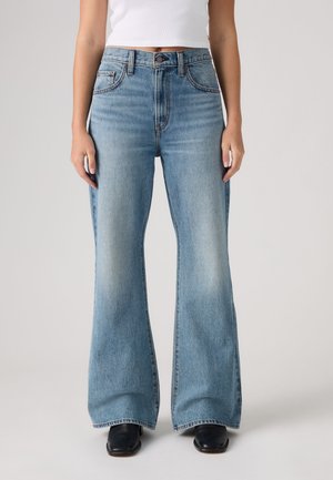 Femme portant un jean évasé taille haute bleu clair avec poches avant et chaussures noires à bout carré, debout sur un fond uni.