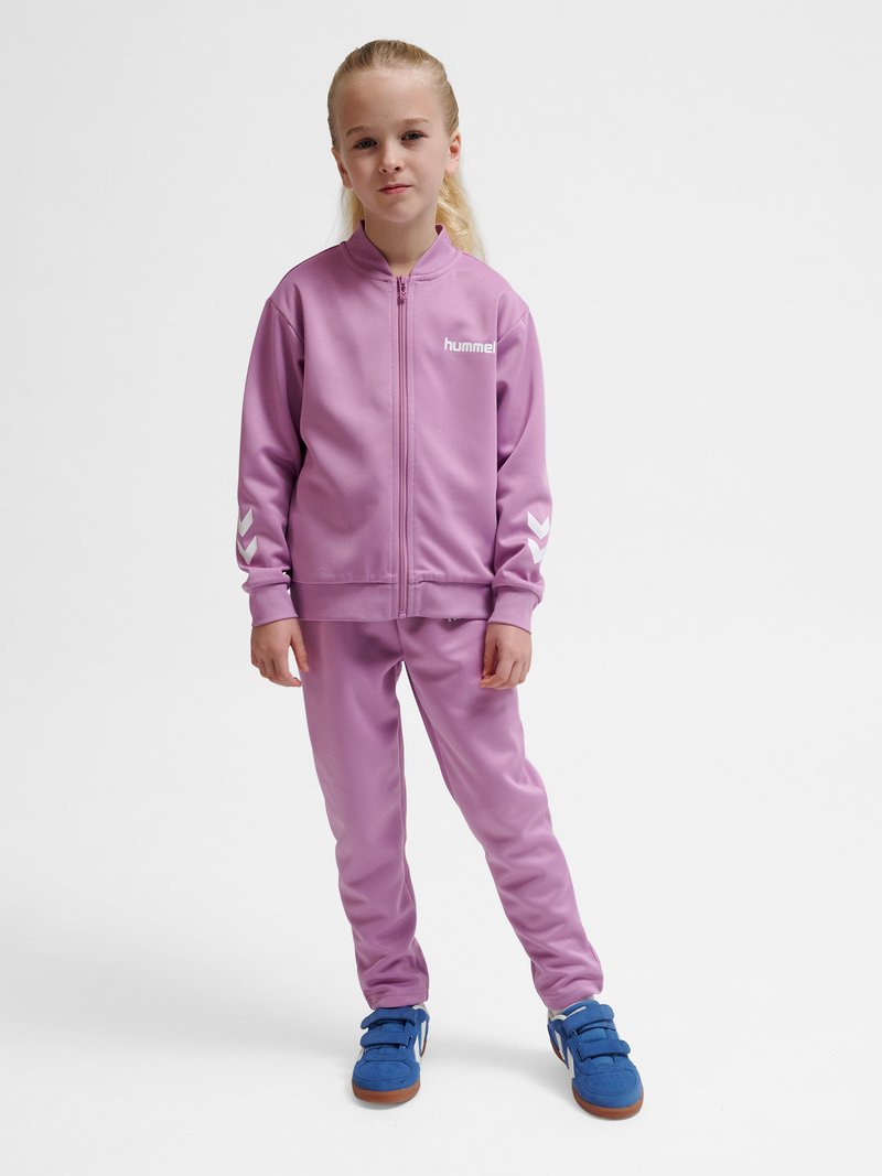 Jeune fille debout en survêtement violet avec des logos blancs et des baskets bleues à scratch, sur un fond blanc uni.