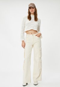 Junge Frau in beigen Cargo-Hosen, einem weißen, cropped Strickpullover, weißen Stiefeln und einer braunen karierten Mütze, die mit einer Hand in der Tasche steht.