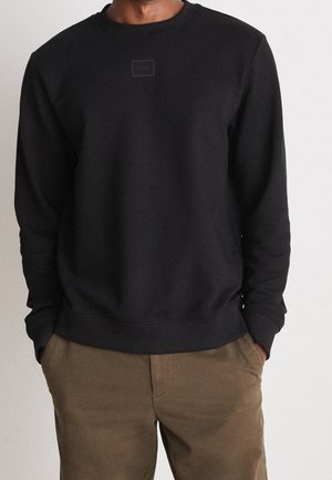 Man wearing black crewneck sweatshirt with small front logo and brown pants, hands casually in pockets, against plain light background.