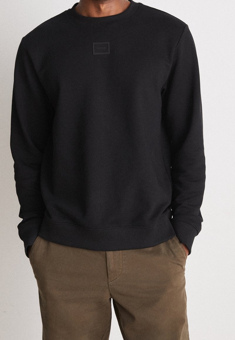 Homme portant un sweat-shirt noir à col rond avec un petit logo sur le devant et un pantalon marron, les mains dans les poches de manière décontractée, sur un fond clair uni.