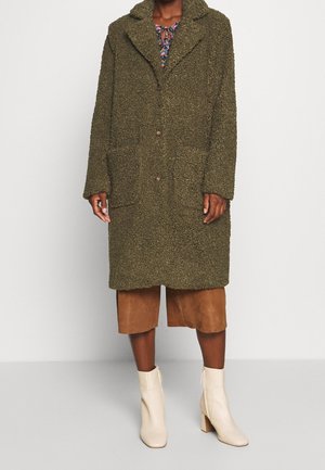 Personne portant un manteau texturé olive vert jusqu'aux genoux, un pantalon marron large et court, et des bottines crème, debout devant un fond blanc uni.