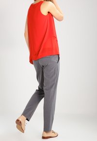 Donna che indossa una blusa rossa senza maniche, pantaloni grigi su misura e mocassini beige, in piedi con un piede leggermente sollevato.