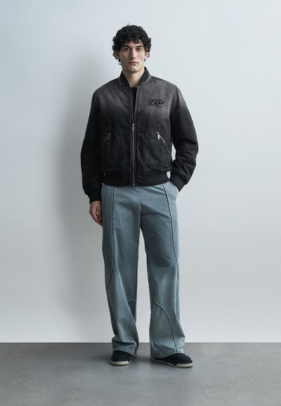 Νεαρός με σγουρά μαλλιά που φοράει μαύρο bomber μπουφάν με διαβάθμιση χρώματος, ανοιχτό μπλε παντελόνι φαρδύ στο πόδι με ραφές και μαύρα αθλητικά παπούτσια, όρθιος.