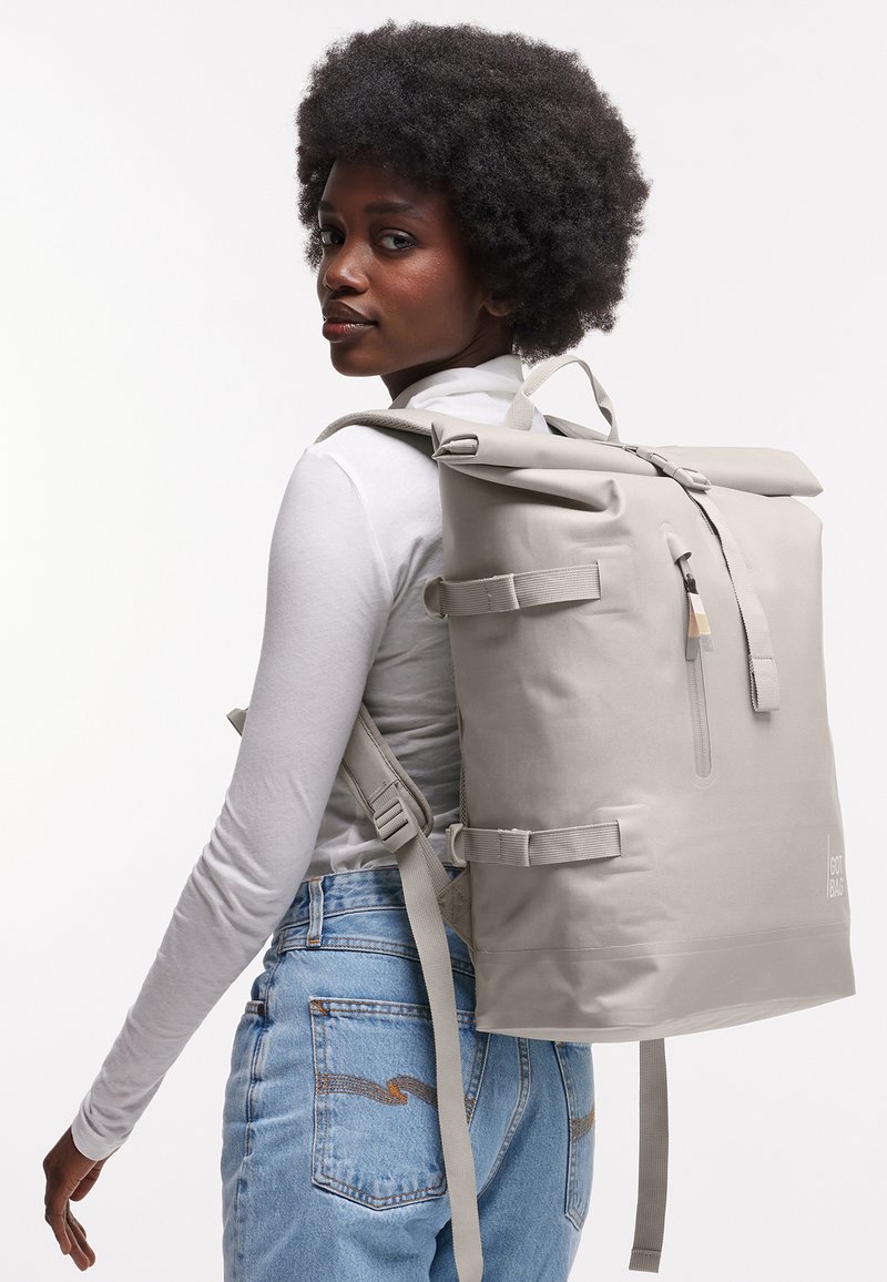 Femme aux cheveux bouclés portant un haut blanc à manches longues et un jean, portant un grand sac à dos gris clair à fermeture roulante dans son dos.