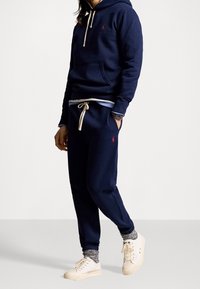 Marinblå huvtröja med dragsnören, framficka; matchande marinblå sweatpants med elastiska muddar; bärs med vita sneakers och mönstrade strumpor.