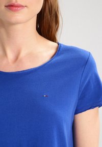 Blå bomullströja med kort ärm och rund halsringning, med en liten logotyp i röd, vit och blå på bröstområdet.