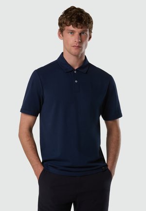 Giovane uomo con capelli ricci che indossa una maglietta polo blu navy a maniche corte con due bottoni, mani in tasca, su sfondo chiaro uniforme.