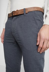 Pantaloni grigi strutturati con tasche frontali, abbinati a una cintura in pelle marrone con fibbia rettangolare, che enfatizzano un design moderno.