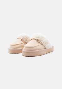 Beige slippers lavet af blødt suede med en fluffy hvid pelskant og gyldne hardware detaljer. Har en tykk, tekstureret sål for ekstra komfort.