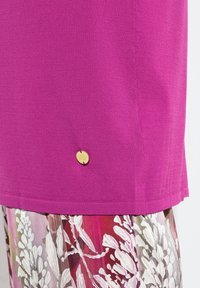 Fialový pletený top s malým kulatým zlatým emblémem u lemu, doplněný květovanou sukní v odstínech bílé, růžové a červené.