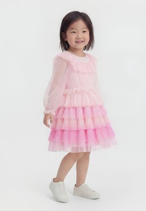 Smilende ung pige iført en lyserød lagdelt tulekjole med lange, gennemsigtige ærmer og hvide sneakers, stående på en hvid baggrund.