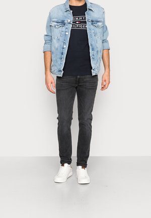 Lys blå denimjakke over en marineblå T-shirt med logo, parret med sorte skinny jeans og hvide sneakers. Pæne syningsdetaljer er synlige.