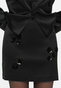 Czarna satynowa sukienka z dużą kokardą w talii oraz ozdobami z czarnych i srebrnych cekinów w kwiatowe wzory wokół dołu.