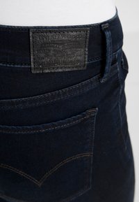 Vaqueros de mezclilla azul oscuro con un parche de cuero con el logo en la cintura, costuras visibles y diseño tradicional de bolsillo trasero.