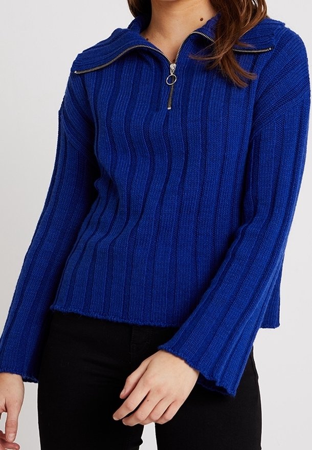 Blå ribbad stickad tröja med halvdold dragkedja, långa utsvängda ärmar och rå fåll. Har en texturerad tyg och glänsande dragkedjedetalj.