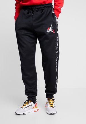 Osoba nosząca czarne spodnie dresowe Air Jordan z logo na udzie i paskiem, białe trampki z żółtymi sznurówkami oraz czerwoną bluzkę.