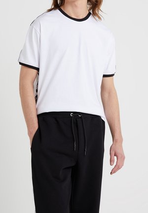 Mand iført hvid t-shirt med korte ærmer og sort kant samt sorte snørede bukser med hænder afslappet ned langs siderne.