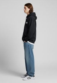 Felpa nera con cappuccio e testo bianco, abbinata a jeans baggy blu chiaro e sneakers bianche con strisce nere. Composizione di vista laterale.