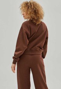 Sweatshirt en coton marron avec une coupe décontractée, des poignets et une taille côtelés. Porté avec un pantalon assorti, montrant une texture douce et un design décontracté.