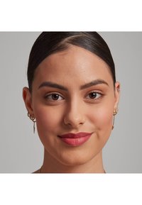Boucles d'oreilles en or avec des accents texturés et des formes allongées. Peau lisse, de teinte moyenne, avec un maquillage naturel mettant en valeur des lèvres mates.