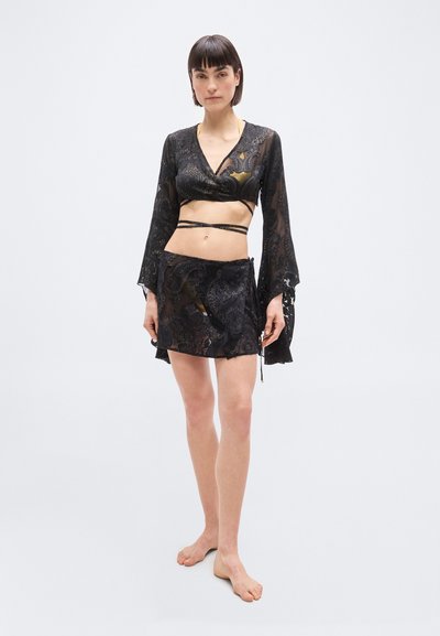 Γυναίκα ξυπόλητη, φορώντας μαύρο, δαντελένιο τοπ τύπου wrap crop με μακριά μανίκια καμπάνα και το αντίστοιχο διάφανο μίνι φούστα πάνω από χρυσό μαγιό.