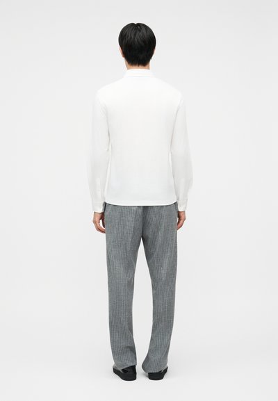 Άνδρας που στέκεται με την πλάτη στραμμένη, φοράει λευκό πουκάμισο με μακριά μανίκια και γκρι καρό παντελόνι με μαύρα παπούτσια, σε ένα απλό φόντο.