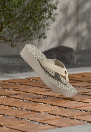Μπεζ thong σανδάλι με σχέδιο στην πελματιαία επιφάνεια, αιωρούμενο πάνω από επιφάνεια από τερακότα πλακάκια και βότσαλα, με θολό φόντο πρασίνου και λίθινης τοιχοποιίας.